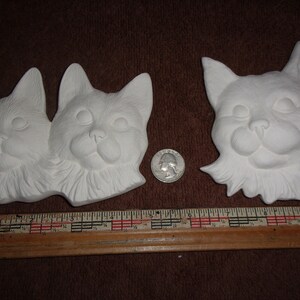 May include: Two unpainted ceramic cat face wall hangings and a single cat face. The cat faces are next to a quarter and a ruler. The cat faces are white and have detailed features.