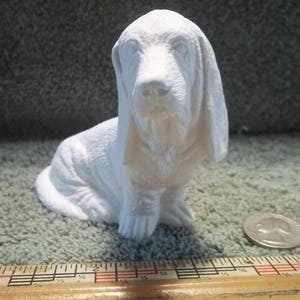May include: A small, white basset hound figurine. The dog is sitting with its head slightly tilted, showcasing its long ears and droopy face. The figurine is on a textured surface, with a ruler and a coin nearby.