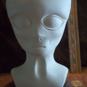 May include: A white alien head sculpture with large, almond-shaped eye sockets and a small mouth. The sculpture has a smooth, matte finish and a slender neck. A ruler and a coin are in the background.