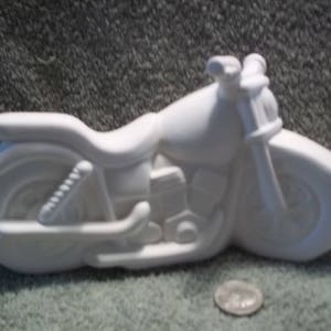 May include: A white ceramic motorcycle figurine with a smooth, unpainted finish. The object features a detailed engine, seat, handlebars, and wheels. A wooden ruler is partially visible in the background, and a silver coin rests in the foreground.