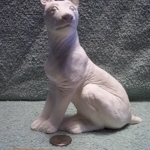 May include: A white ceramic dog figurine in a seated position, featuring detailed facial features and textured fur. A wooden ruler is placed in the foreground, showing the figurine stands approximately 6 inches tall next to a small metal coin.