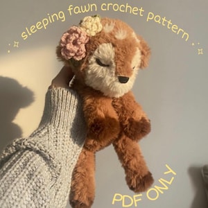 May include: A handmade brown and cream-coloured fawn plushie with closed eyes, adorned with a floral crown. The text "sleeping fawn crochet pattern" is arched above the fawn. The words "PDF ONLY" are written in a circle below the fawn.