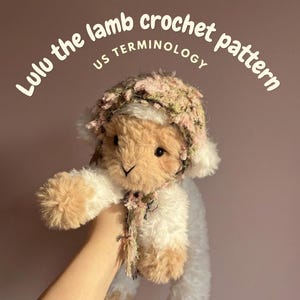 Op de afbeelding: Een pluche lammetjesspeelgoed met een gehaakte muts en bloemaccenten. Het lam wordt tegen een roze achtergrond gehouden. Tekst op de afbeelding luidt "Lulu the lamb crochet pattern US terminology". Het lam heeft bruin en wit bont.