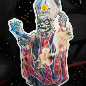 May include: Holographic sticker featuring a skeletal figure in a red and black robe with crosses, a tall white hat, and a smiley face emblem. The figure is holding a glowing orb. The sticker has a reflective, iridescent finish.