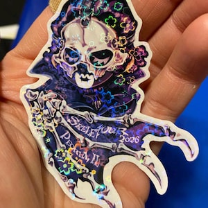 May include: Holographic sticker with a skull design, floral details, and a skeleton figure. The text "SKELE YOUR PEORIA, II 2026" is displayed. The sticker has a dark purple and iridescent colour scheme.