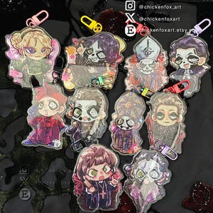 以下が含まれることがあります： 様々なフェイスペイントデザイン、カラフルな留め具、グリッターのアクセントが施された、ちびキャラ風のイラストが描かれた10個のアクリルキーホルダーのコレクション。右上に@chickenfox_artというテキストが表示されています。