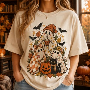 Puede incluir: Una camiseta de color crema con un estampado gráfico de tres fantasmas, uno con un gorro naranja y una taza, otro con un patrón de cuadros y un lazo, y un tercero con una corona de flores, junto a un gato negro, una calabaza y murciélagos.