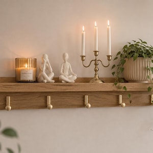 May include: A light oak wood wall shelf featuring five gold-toned metal hooks along the bottom edge. The shelf surface holds a glass candle jar, two white seated human figurines, a three-arm brass candelabra with white candles, and a potted plant.