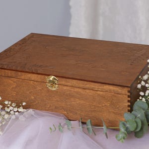 Op de afbeelding: Een houten doos met een scharnierend deksel en een messing sluiting. De doos is donkerbruin gebeitst en heeft een houtnerfpatroon. De doos staat op een wit oppervlak.