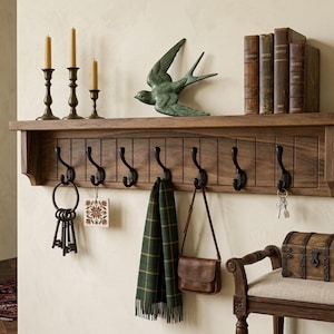 May include: A wooden coat rack with six black hooks, holding keys, a scarf, and a purse. Above, a shelf displays books, candles, and a bronze bird sculpture. The rack is made of dark wood, creating a rustic, vintage aesthetic.