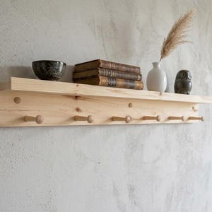 May include: A light wood wall shelf with a top shelf and a lower shelf with pegs. The top shelf holds a black bowl, books, a white vase with a plant, and a small owl figurine. The shelf is mounted on a light-coloured wall.
