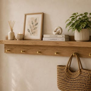 Puede incluir: Una estantería de pared de madera con ganchos de latón, que sostiene una impresión botánica enmarcada, libros y una planta en maceta. Una bolsa de mano tejida cuelga debajo. Incluye un jarrón con cañas y una vela.