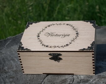 Può includere: Una scatola di legno color chiaro con angoli decorativi in metallo e una chiusura. Il coperchio presenta il nome "Victorija" in corsivo all'interno di una ghirlanda floreale. La scatola è posta su uno sfondo verde sfocato.