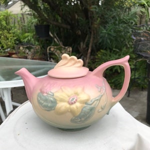 May include: A pink and yellow ceramic teapot with a floral design. The teapot has a lid with a decorative knob and a spout on the side. The teapot is sitting on a white surface.