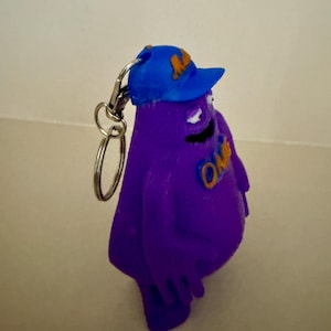 May include: Purple 3D printed keychain of a cartoon character wearing a blue baseball cap with the letter "L" on it. The character has a big smile and is wearing a shirt that says "OMG".