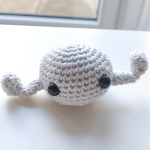 May include: A small, crocheted, gray toy with two black eyes and two small arms. The toy is sitting on a white surface.