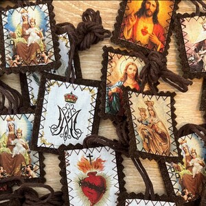 Pode incluir: Uma coleção de cartões de oração religiosos com imagens de Jesus, Maria e outras figuras religiosas. Os cartões são retangulares com uma borda decorativa e presos a um cordão marrom. As imagens são coloridas e detalhadas.