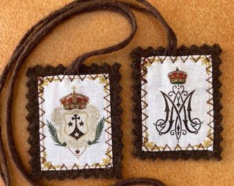 May include: Two square religious pendants with brown trim and cord. One features a crown over a shield with a cross and olive branches. The other has a crown over a stylized "M". Both are on a tan background.