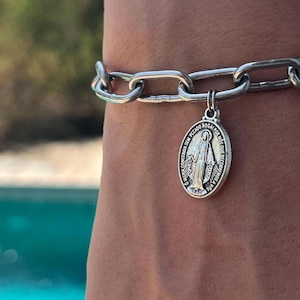 Può includere: Bracciale a catena argentata con un ciondolo circolare. Il ciondolo presenta l'immagine di una figura religiosa e del testo. Il bracciale è composto da grandi maglie ad incastro. Lo sfondo è sfocato, con una piscina turchese visibile.