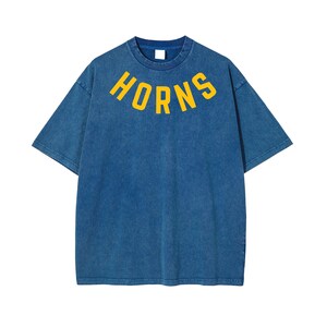 May include: Blue t-shirt with the word "HORNS" in yellow arched across the chest. The shirt has short sleeves and a crew neck. The fabric has a faded, vintage look.