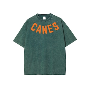 May include: Green t-shirt with the word "CANES" in orange block letters across the chest. The shirt has a vintage, washed appearance and a crew neck. The short-sleeved shirt is made of a soft material.