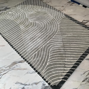 May include: A gray and white patterned rug with a black border. The rug has a wavy design and is made of a soft, plush material.