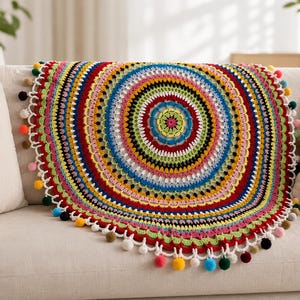 Puede incluir: Una colorida manta circular de ganchillo con un vibrante diseño de mandala. La manta presenta anillos concéntricos de color rojo, amarillo, verde, azul y negro. El borde está adornado con festones blancos y pompones de colores. La manta está sobre un sofá beige.