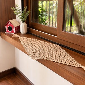 May include: A beige crocheted table runner with a diamond-shaped pattern and a tassel at each pointed end. The runner features an open-work design and is crafted from a light-colored yarn, displaying a consistent texture throughout the piece.