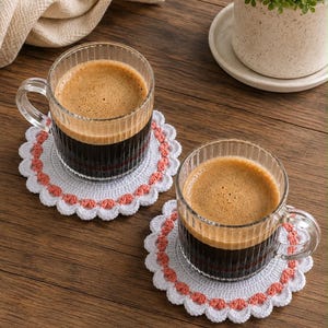 Pode incluir: Duas canecas de vidro transparente com estrias verticais cheias de café castanho escuro, cada uma sobre um porta-copos redondo de croché branco e cor de pêssego com borda festonada. Os porta-copos têm um centro branco e uma borda decorativa.