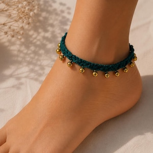 May include: A dark teal crocheted anklet featuring a series of small, round, metallic gold-toned jingle bells spaced evenly along the length of the band. The anklet is constructed from a textured yarn material and secures around the ankle.