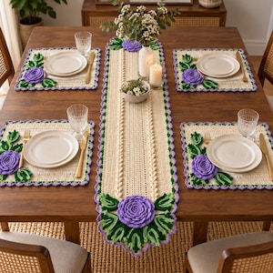 May include: A cream-coloured crochet table runner and four matching square placemats, each featuring three-dimensional purple rose appliqués with green leaves and a scalloped purple border, arranged on a wooden dining table with white plates.