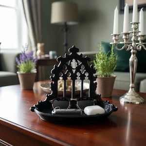May include: A black metal tray featuring an ornate, arched architectural design reminiscent of a cathedral window. The tray holds a pair of black-rimmed sunglasses, a car key fob, and a white wireless earbud charging case on its base.