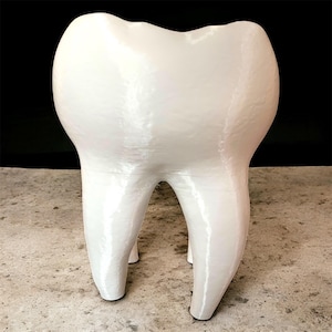 Może przedstawiać: Duży, biały, dekoracyjny przedmiot w kształcie zęba. Rzeźba ma błyszczące wykończenie i jest podparta trzema nogami w kształcie korzeni. Dzieło sztuki o tematyce dentystycznej jest umieszczone na neutralnym tle.