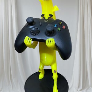 May include: A yellow banana-shaped figurine holding a black video game controller. The figurine has brown details and is standing on a black circular base.