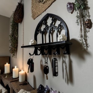 Puede incluir: Llavero de metal negro con diseño de arco gótico. El llavero tiene un estante con dos figuras de calaveras blancas y ganchos para llaves. El llavero está montado en una pared gris claro. Las llaves cuelgan de los ganchos.