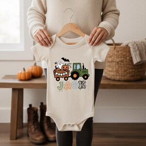 Puede incluir: Un body de bebé de manga corta en color crema con un gráfico de un tractor verde tirando de un remolque lleno de calabazas, un fantasma y un murciélago. Debajo del remolque, el nombre JACK está impreso en letras grandes decoradas con telarañas.