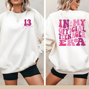 May include: White sweatshirt with pink lettering on the front that says "13 Sophia" and pink lettering on the back that says "In My Official Teenager Era".
