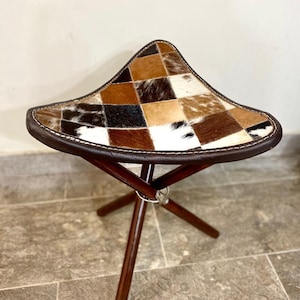 May include: A three-legged stool with a triangular seat. The seat is made of a patchwork of brown, black, and white animal hide. The edges are trimmed with brown leather and white stitching. The legs are dark brown wood.