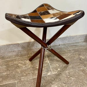 May include: A three-legged stool with a seat made of a patchwork of brown, tan, and white cowhide. The seat is edged with dark brown leather. The legs are dark brown wood, joined with a metal ring.