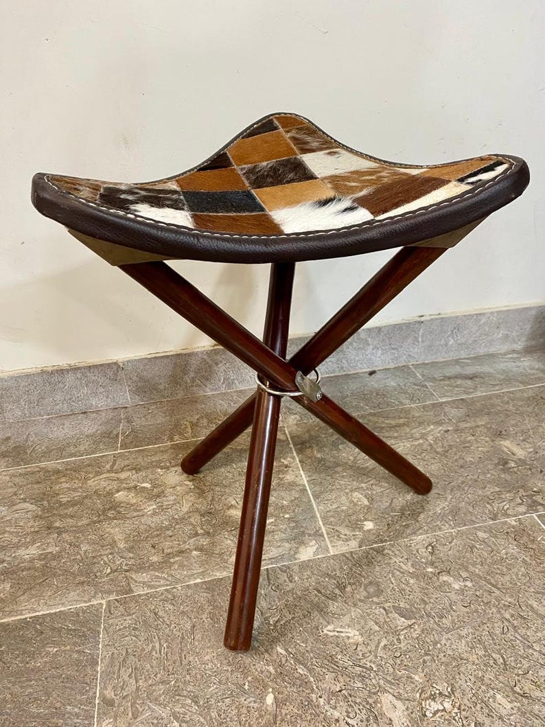 May include: A three-legged stool with a seat made of a patchwork of brown, tan, and white cowhide. The seat is edged with dark brown leather. The legs are dark brown wood, joined with a metal ring.