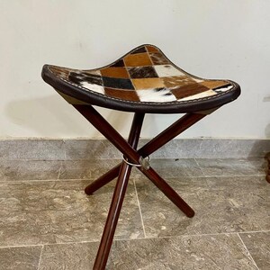 May include: A folding stool featuring a patchwork cowhide seat in brown, tan, and white. The seat is trimmed with dark brown leather and stitched with white thread. The dark wood legs are joined at the centre with a metal hinge.