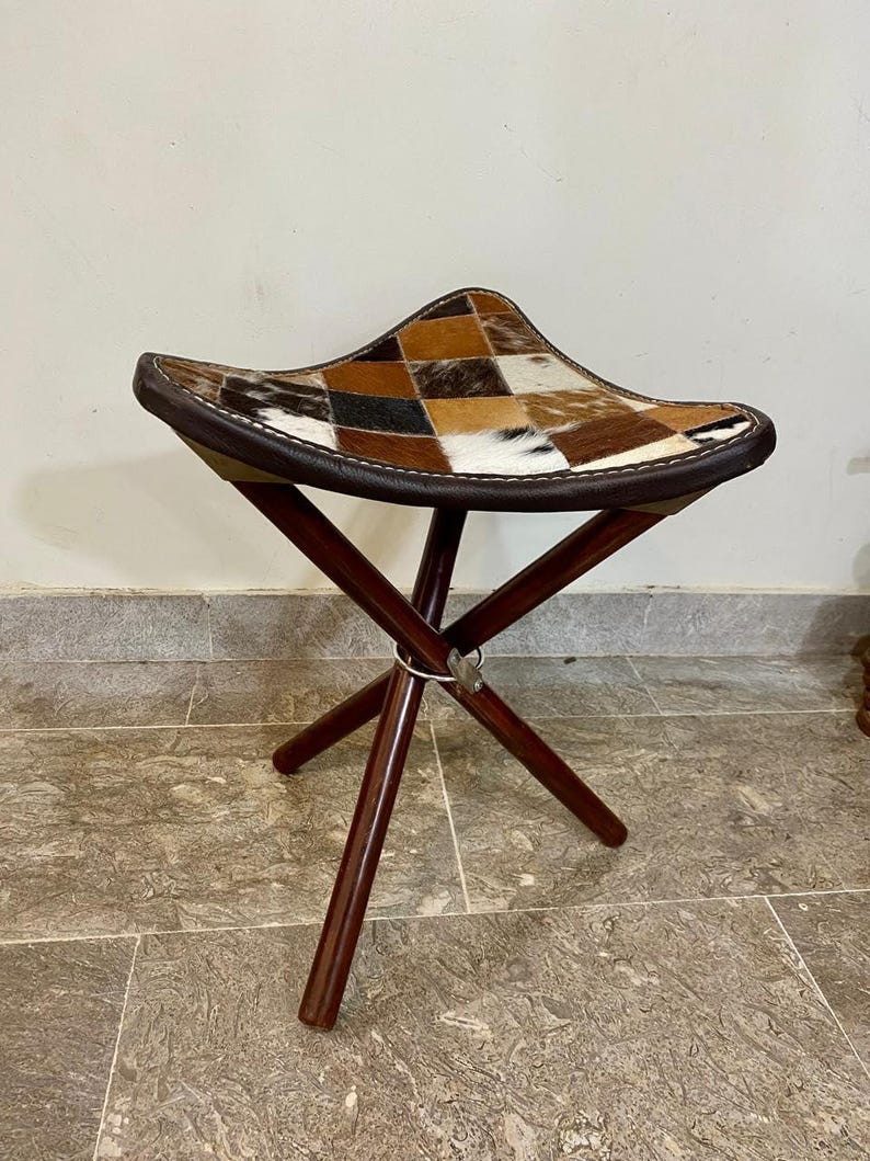 May include: A folding stool featuring a patchwork cowhide seat in brown, tan, and white. The seat is trimmed with dark brown leather and stitched with white thread. The dark wood legs are joined at the centre with a metal hinge.