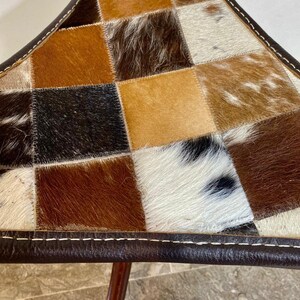 May include: A triangular stool with a patchwork design. The seat is made of squares of cowhide in shades of brown, tan, and white. The edges are trimmed with dark brown leather and white stitching. The legs are made of dark wood.