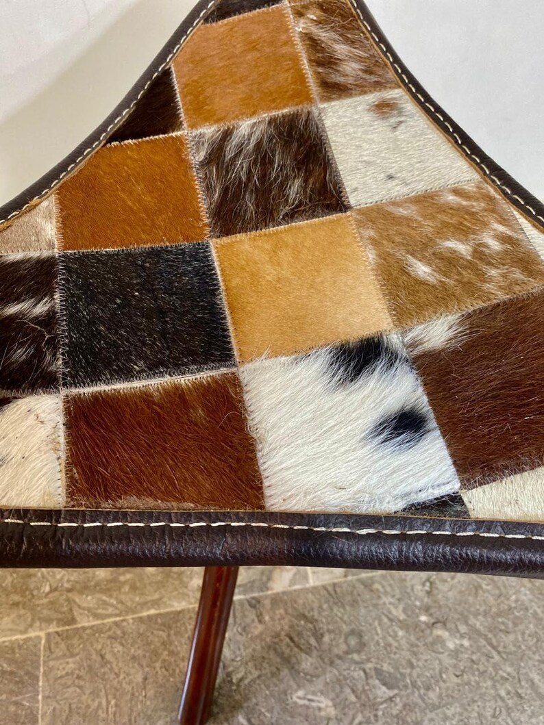 May include: A triangular stool with a patchwork design. The seat is made of squares of cowhide in shades of brown, tan, and white. The edges are trimmed with dark brown leather and white stitching. The legs are made of dark wood.