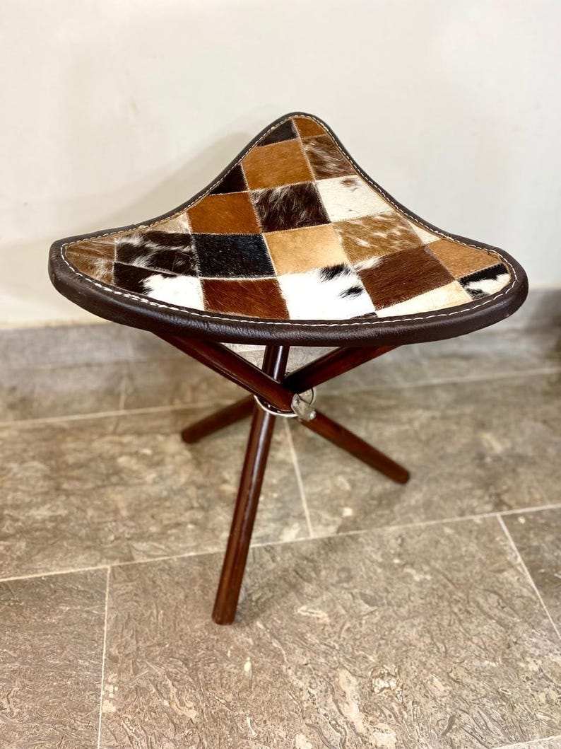 May include: A three-legged stool with a triangular seat. The seat is a patchwork of brown, black, and white animal hide. The edges are trimmed with brown leather and white stitching. The legs are dark brown wood.