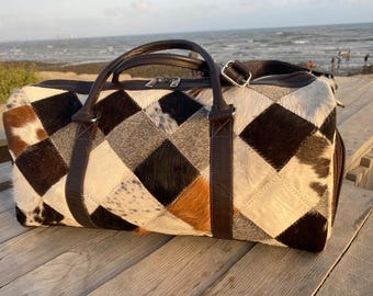 May include: A duffel bag featuring a patchwork design of cowhide squares in shades of black, white, brown, and tan. The bag includes dark brown leather handles, a matching adjustable shoulder strap, and a top zipper closure with silver hardware.