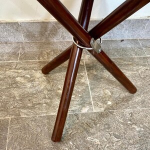 May include: A dark brown wooden folding table with a circular metal ring and metal bracket. The table legs are angled and taper towards the floor. The table is on a tiled surface.