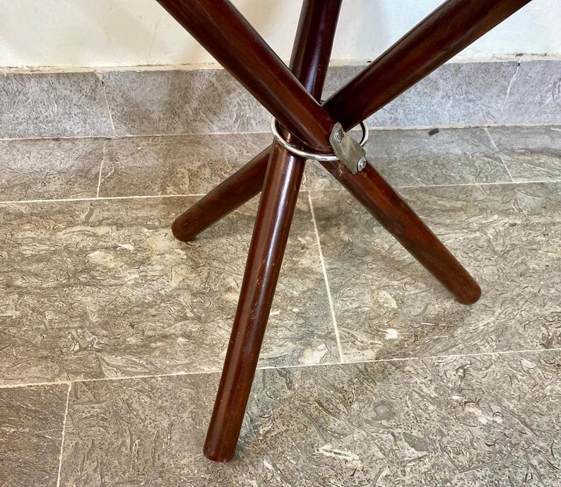 May include: A dark brown wooden folding table with a circular metal ring and metal bracket. The table legs are angled and taper towards the floor. The table is on a tiled surface.