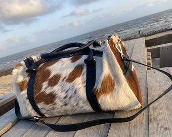 May include: A duffel bag featuring a brown and white cowhide pattern with black leather trim, black handles, and a detachable black shoulder strap. The bag has a zippered closure and metal hardware, showcasing a textured hair-on-hide exterior.