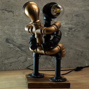 Può includere: Una lampada da tavolo in stile steampunk in oro e nero realizzata con tubi. La lampada ha due lampadine e una base in legno.